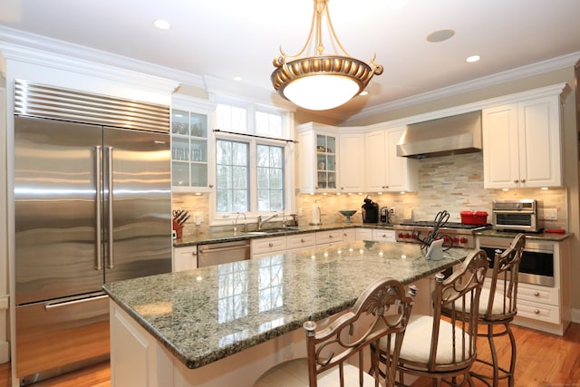 kitchen with wall chimney range hood, dark stone countertops, stainless steel appliances, a kitchen island, and decorative light fixtures