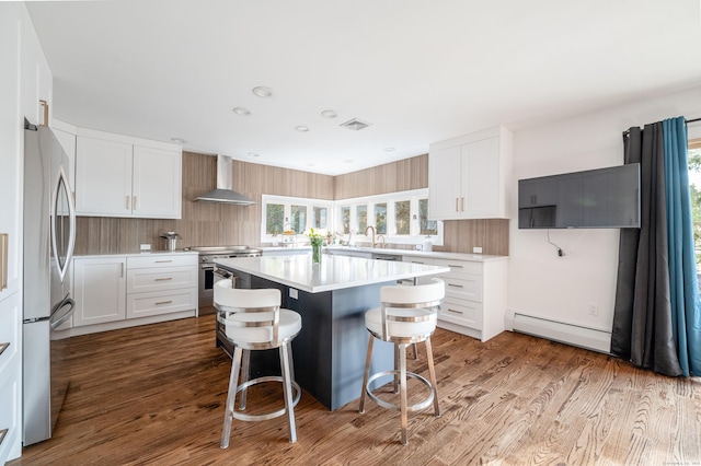 kitchen with wall chimney range hood, appliances with stainless steel finishes, a baseboard heating unit, a center island, and white cabinets