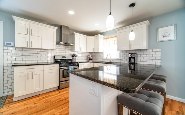 kitchen featuring decorative light fixtures, white cabinets, a kitchen bar, gas range, and wall chimney range hood