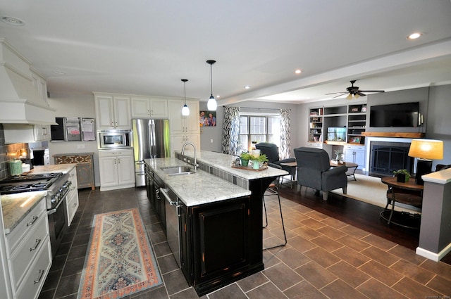 kitchen featuring a kitchen island with sink, stainless steel appliances, a fireplace, a sink, and white cabinetry