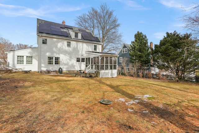 back of house featuring a sunroom, roof mounted solar panels, a chimney, and a lawn