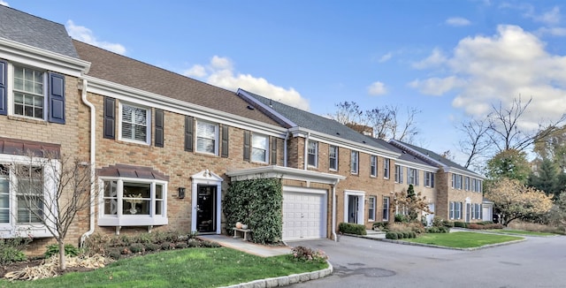 townhome / multi-family property featuring driveway, a garage, a shingled roof, brick siding, and a front yard