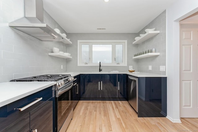 kitchen featuring appliances with stainless steel finishes, blue cabinets, sink, light wood-type flooring, and wall chimney exhaust hood