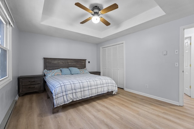 bedroom with light hardwood / wood-style flooring, ceiling fan, a tray ceiling, a baseboard radiator, and a closet