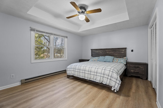 bedroom with ceiling fan, baseboard heating, hardwood / wood-style floors, a tray ceiling, and a closet