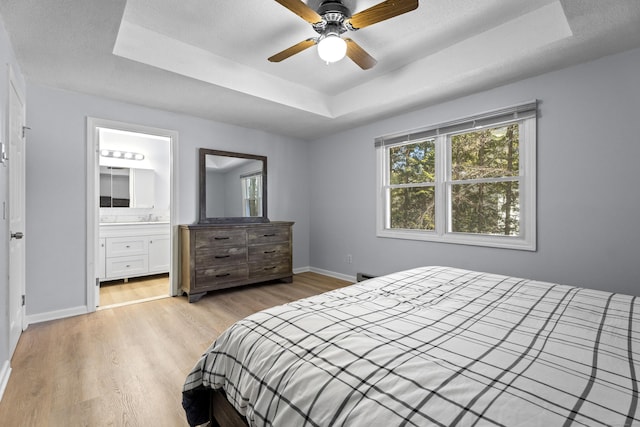 bedroom with ceiling fan, connected bathroom, a tray ceiling, light hardwood / wood-style floors, and a textured ceiling