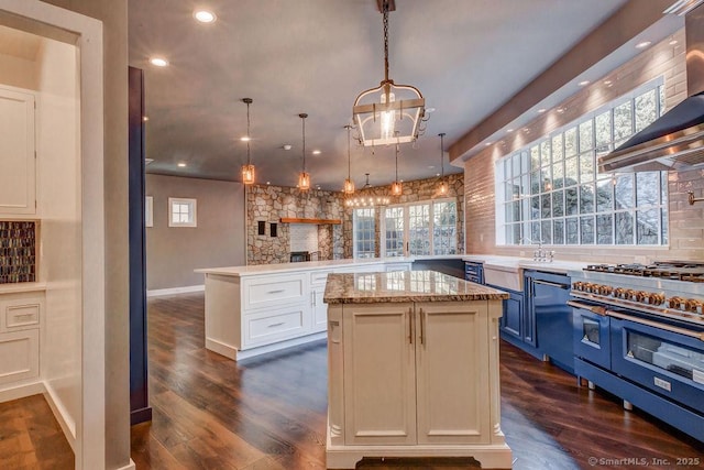 kitchen with wall chimney range hood, double oven range, hanging light fixtures, a center island, and light stone counters