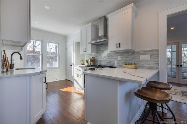 kitchen with wall chimney range hood, sink, a kitchen breakfast bar, ornamental molding, and white cabinets