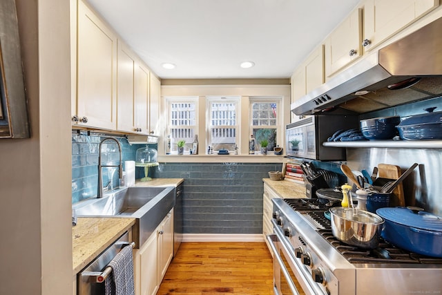 kitchen with a sink, light wood finished floors, appliances with stainless steel finishes, under cabinet range hood, and baseboards