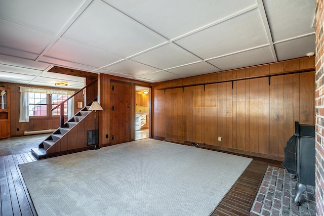 unfurnished living room with stairway, wood finished floors, wood walls, a wood stove, and a baseboard radiator