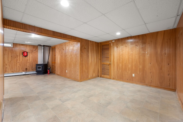 basement featuring wood walls, a drop ceiling, a wood stove, and a baseboard heating unit