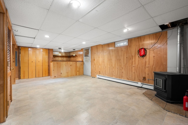 basement with baseboard heating, a paneled ceiling, a wood stove, and wood walls