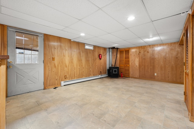 basement featuring a paneled ceiling, a wood stove, wood walls, and baseboard heating