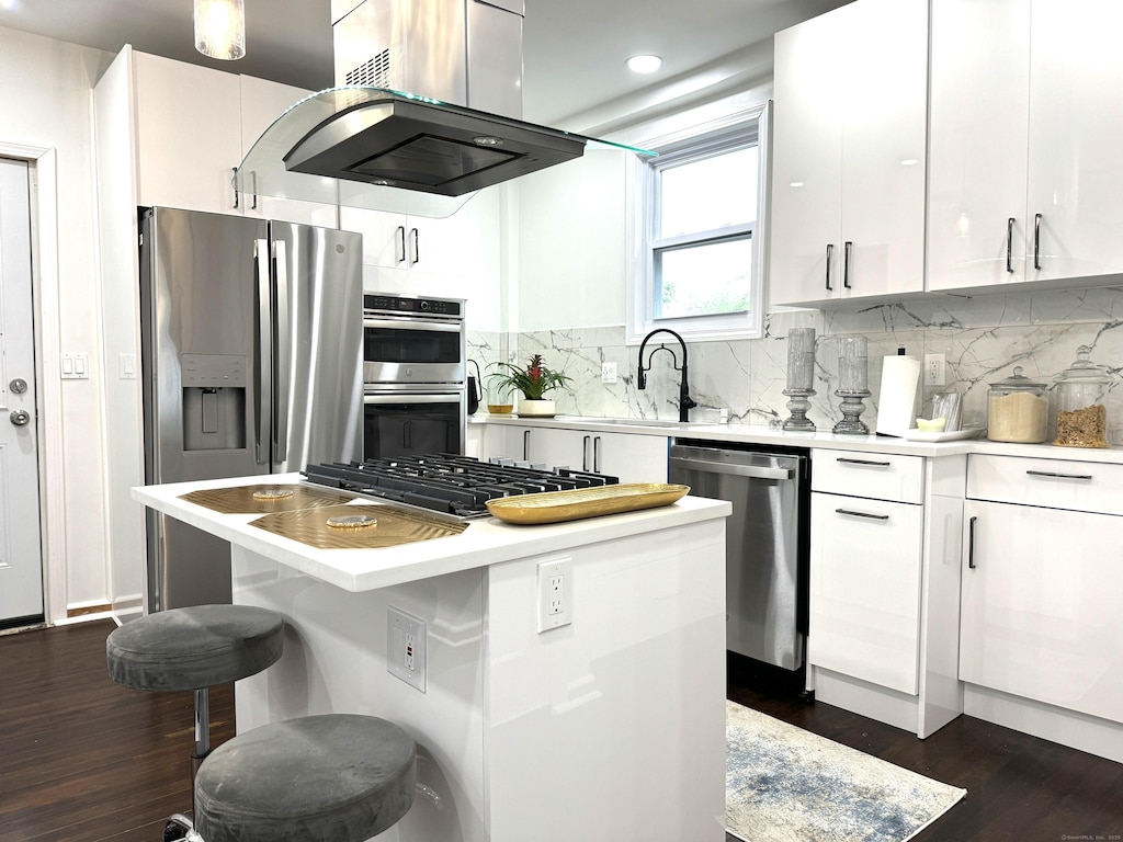 kitchen with stainless steel appliances, island range hood, white cabinets, a kitchen island, and decorative backsplash