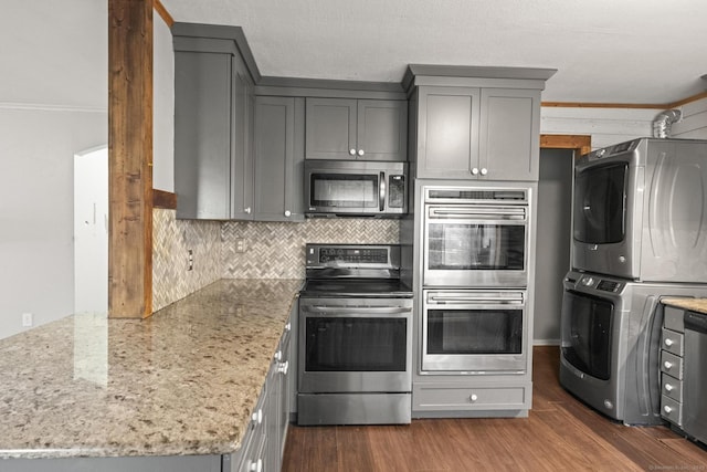 kitchen with gray cabinets, appliances with stainless steel finishes, stacked washer / dryer, light stone counters, and dark wood-type flooring