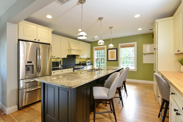 kitchen with an island with sink, appliances with stainless steel finishes, hanging light fixtures, light stone countertops, and custom exhaust hood