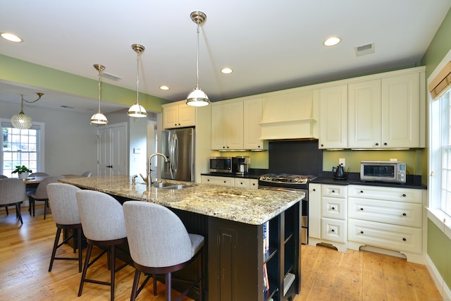 kitchen featuring a center island with sink, visible vents, hanging light fixtures, stainless steel appliances, and premium range hood