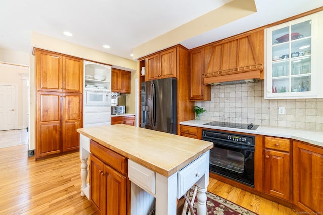 kitchen with light wood-type flooring, decorative backsplash, black appliances, brown cabinetry, and custom range hood