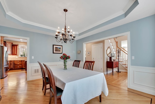 dining area with light wood-type flooring, stairway, a raised ceiling, and a wainscoted wall