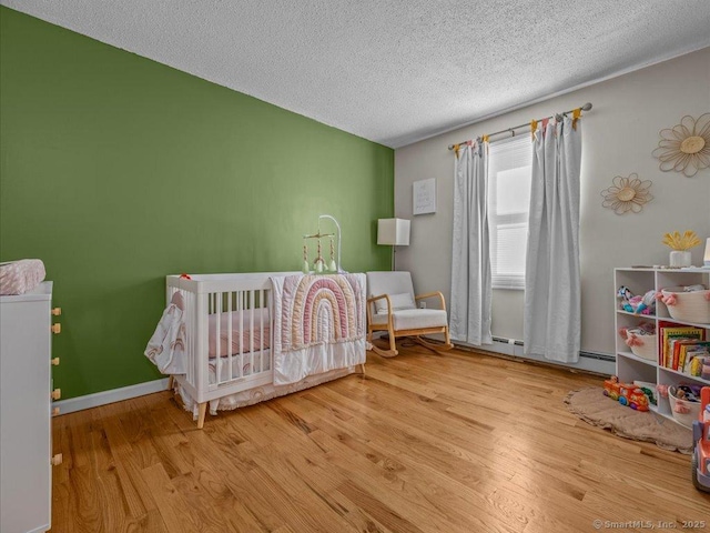 bedroom with a crib, a textured ceiling, baseboard heating, and light hardwood / wood-style flooring