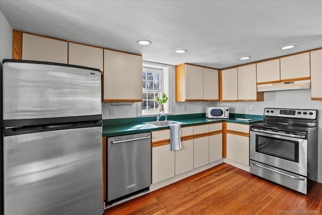 kitchen featuring under cabinet range hood, light wood-type flooring, appliances with stainless steel finishes, cream cabinets, and a sink