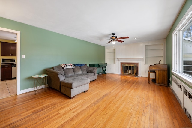 living room featuring a brick fireplace, light wood-type flooring, radiator, built in features, and ceiling fan