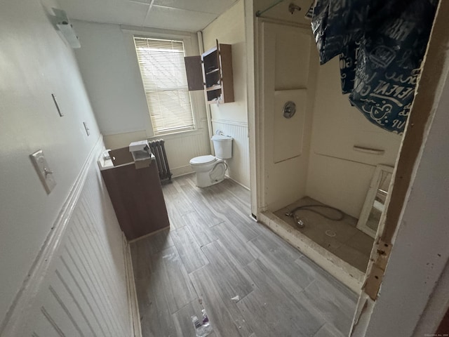 bathroom featuring a shower, a drop ceiling, toilet, a wainscoted wall, and wood finished floors