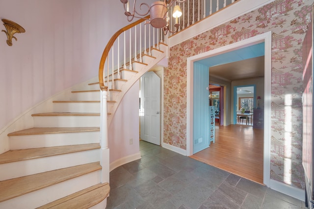 staircase featuring an inviting chandelier, ornamental molding, stone finish flooring, baseboards, and wallpapered walls