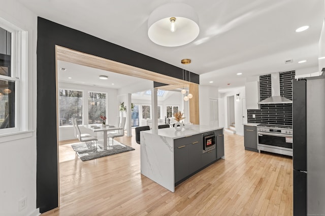 kitchen featuring wall chimney exhaust hood, light wood-type flooring, appliances with stainless steel finishes, pendant lighting, and backsplash