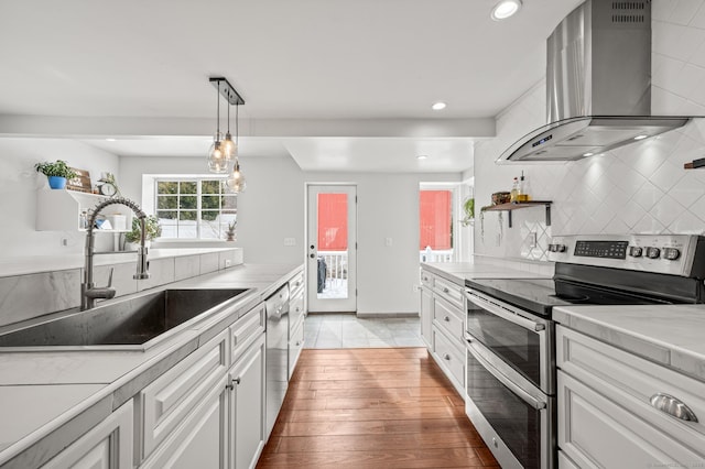 kitchen featuring stainless steel appliances, wall chimney range hood, decorative light fixtures, sink, and white cabinetry