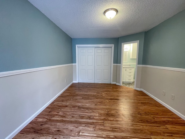 unfurnished bedroom with a closet, connected bathroom, a textured ceiling, wood finished floors, and baseboards