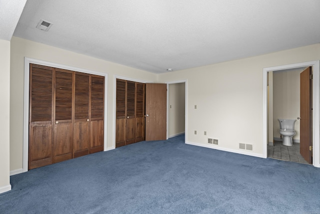 unfurnished bedroom featuring multiple closets, ensuite bath, a textured ceiling, and dark colored carpet