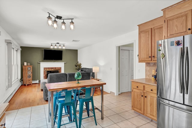 kitchen featuring light tile patterned floors, stainless steel refrigerator, baseboard heating, tasteful backsplash, and a brick fireplace