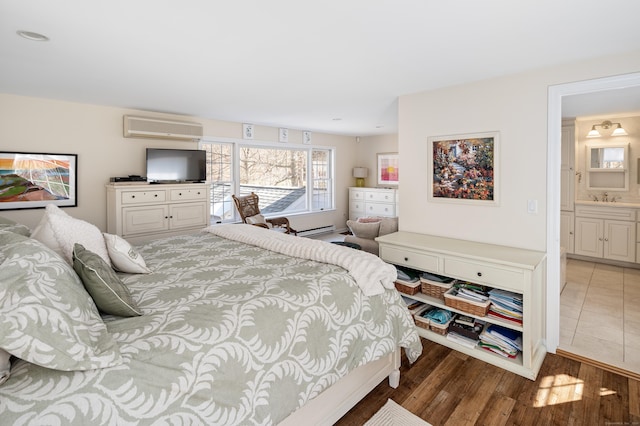 bedroom with a baseboard heating unit, wood finished floors, ensuite bath, a wall mounted AC, and a sink