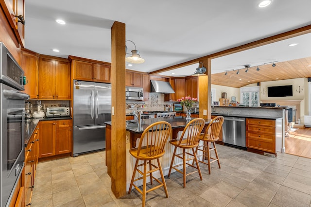 kitchen with decorative backsplash, appliances with stainless steel finishes, brown cabinets, open floor plan, and wall chimney range hood