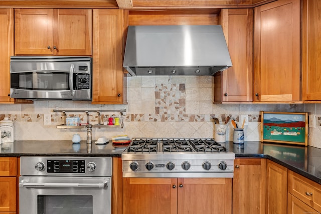 kitchen featuring stainless steel appliances, decorative backsplash, wall chimney exhaust hood, brown cabinetry, and dark stone countertops