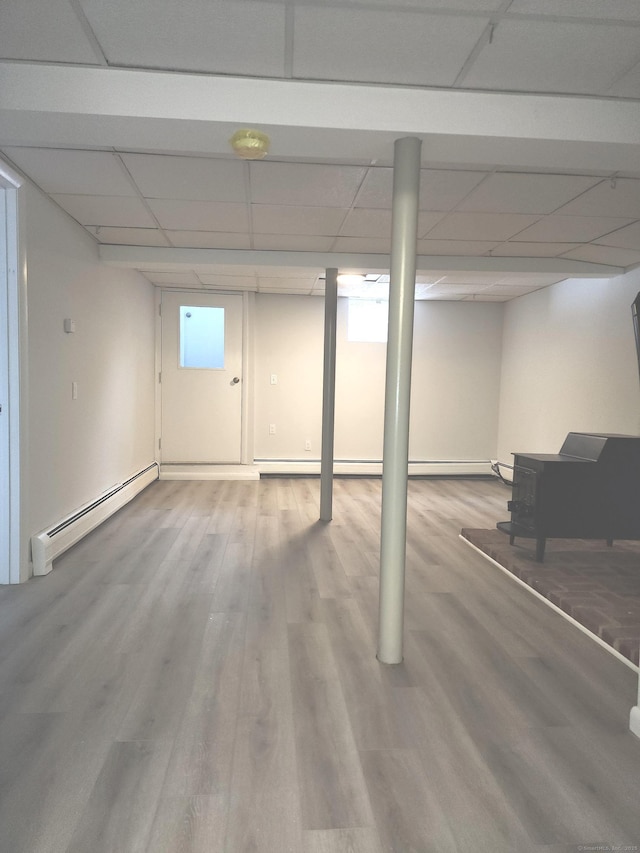 below grade area featuring baseboard heating, a wood stove, wood finished floors, and a paneled ceiling