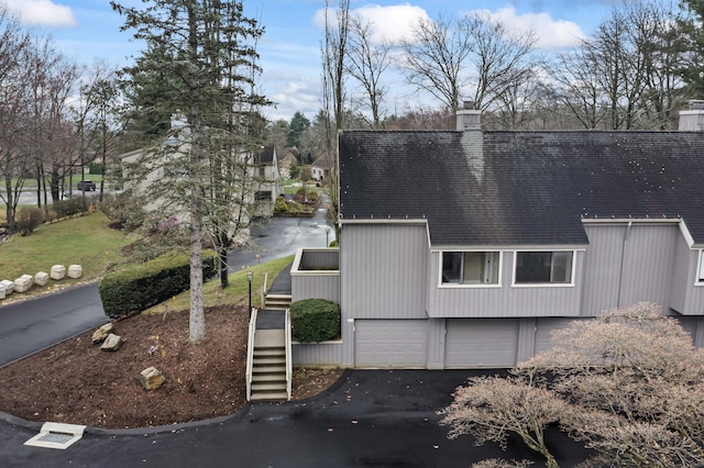 view of side of home featuring roof with shingles, a chimney, an attached garage, stairs, and aphalt driveway