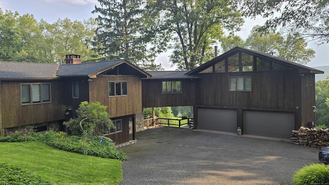 chalet / cabin featuring driveway, a garage, a shingled roof, a chimney, and a front lawn