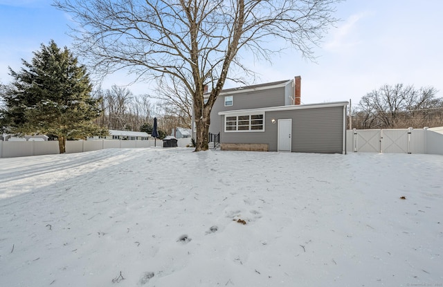 snow covered rear of property featuring a chimney, fence, and a gate