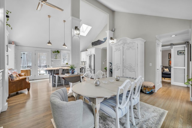 dining space with ceiling fan, high vaulted ceiling, a barn door, a skylight, and light wood-type flooring