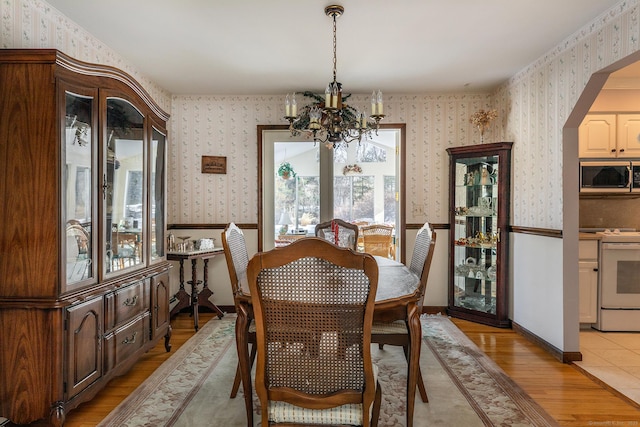 dining room with wallpapered walls, light wood finished floors, arched walkways, a wainscoted wall, and an inviting chandelier