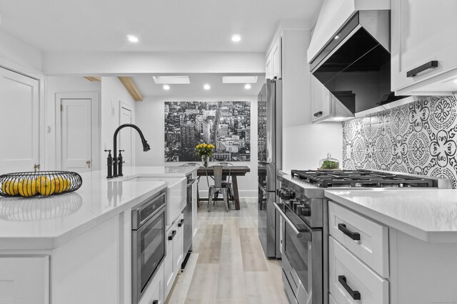 kitchen featuring white cabinets, appliances with stainless steel finishes, light wood-type flooring, wall chimney exhaust hood, and tasteful backsplash