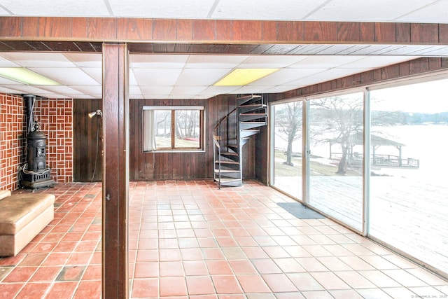 unfurnished living room with a wealth of natural light, a wood stove, a drop ceiling, and wooden walls