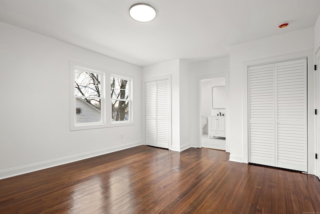 unfurnished bedroom with dark wood-style floors, ensuite bath, two closets, and baseboards