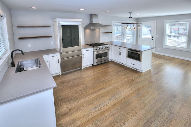 kitchen featuring high end stainless steel range oven, wall chimney range hood, decorative light fixtures, sink, and white cabinetry