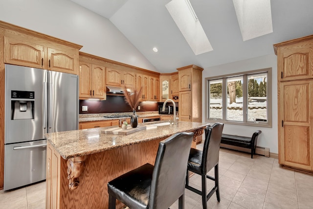 kitchen featuring vaulted ceiling with skylight, a kitchen island with sink, stainless steel appliances, a kitchen breakfast bar, and light stone countertops