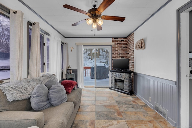 living room with a fireplace, visible vents, a ceiling fan, ornamental molding, and wainscoting