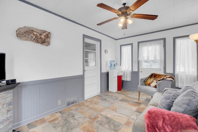 living area with visible vents, wainscoting, ceiling fan, stone finish flooring, and crown molding