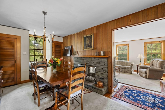 dining space featuring light colored carpet, wooden walls, a stone fireplace, a chandelier, and baseboards